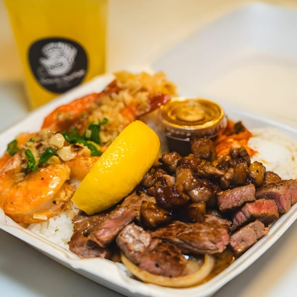 Hawaiian plate lunch with garlic shrimp, steak, rice, and a lemon wedge, with a drink in the background.